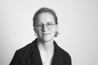 Schwarz-weiße Porträtaufnahme einer Person mit Brille, Ohrringen und zurückgebundenem Haar, die leicht lächelt.