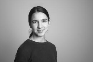 Porträt einer lächelnden Person mit dunklen Haaren, die zu einem Zopf gebunden sind, vor einfarbigem Hintergrund