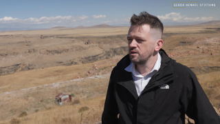 Person mit kurzem Haar und Bart in schwarzer Jacke steht vor einer weiten, trockenen Landschaft mit Hügeln.