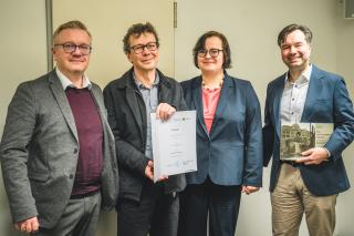 Vier Personen stehen nebeneinander, eine hält eine Urkunde, eine andere ein Buch mit einem historischen Gebäude auf dem Einband, alle tragen formelle Kleidung.