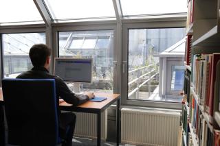 Person sitzt an einem Schreibtisch vor einem Computer in einem Raum mit großen Fenstern und Blick auf ein Dach mit Glasflächen.