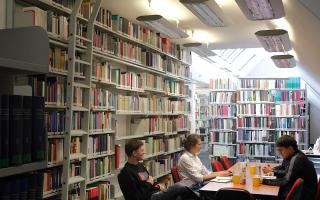 Vier Personen sitzen an einem Tisch in einem schmalen, hohen Raum mit Bücherregalen an den Wänden und Dachfenstern.