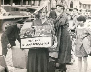 Schwarz-weiße Aufnahme einer Schallplattenverkäuferin in Stockholm der 1930er Jahre mit einem Schild 'DEN NYA GRAMMOFONSKIVAN' und mehreren Schallplatten in der Hand, im Hintergrund Passanten und Autos.