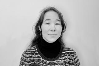 Person mit langen, gewellten Haaren trägt einen gemusterten Pullover über einem schwarzen Rollkragenpullover vor einfarbigem Hintergrund.