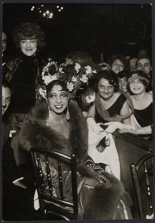 Josephine Baker sitzt elegant in einem Stuhl, trägt ein aufwändiges Kopfschmuck mit Blumen, ein langes Kleid und einen Pelz, umgeben von mehreren Personen in Abendkleidung.