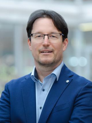 Mann mit Brille, dunklem Haar und blauem Sakko vor unscharfem Hintergrund.