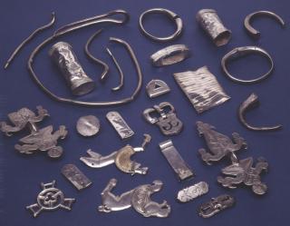 Eine Anordnung von verschiedenen silbernen Schmuckstücken, darunter Ringe, Anhänger, und verzierte Platten, auf einem blauen Untergrund.
