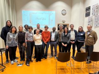 Skupinová fotografie účastníků a účastnic odborného setkání stojících v přednáškové místnosti. Za nimi je promítací plátno se snímkem „Thank You“, na stěně hodiny a informační panely. Lidé stojí v řadě, někteří s jmenovkami, v neformálním pracovním oblečení. Před nimi jsou prázdné židle.