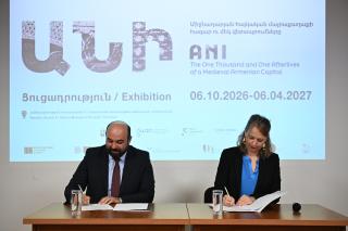Zwei Personen sitzen an einem Tisch und unterschreiben Dokumente vor einer Wand mit einer Projektion zum Thema 'ANI The One Thousand and One Afterlives of a Medieval Armenian Capital' mit Ausstellungsdaten.