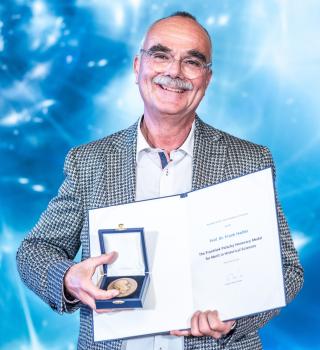 Můž stojící před modrým pozadím. Před sebou drží otevřené desky s diplomem a otevřenou krabičku s medailí.