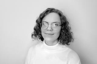 Person mit lockigem, schulterlangem Haar trägt einen weißen Rollkragenpullover vor hellem Hintergrund.