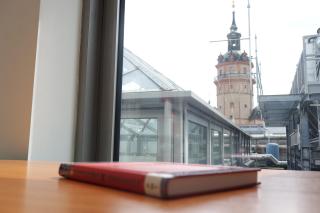 Ein Buch liegt auf einem Tisch an einer Fensterfront, hinter der man den Kirchturm der Nikolaikirche hervorragen sieht.