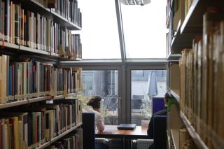 Am Ende eines Gangs mit Bücherregalen auf beiden Seiten sitzen zwei Personen an einer Fensterfront an Tischen und arbeiten am Laptop bzw. lesen in einem Buch.