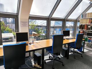 Zu sehen sind drei Arbeitsplätze in der Bibliothek vor einer Fensterfront mit blauen Bürostühlen. Zwei der Arbeitsplätze haben einen Desktop-PC, einer eine Tischlampe. Rechts am Bildrand ist ein Bücherregal zu sehen. Durch das Fenster sieht man einige Pflanzen auf der Dachterasse.