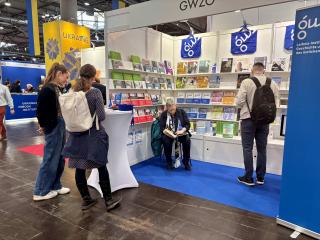 Messestand mit Büchern und Flyern, eine sitzende Person liest, mehrere Personen stehen davor.
