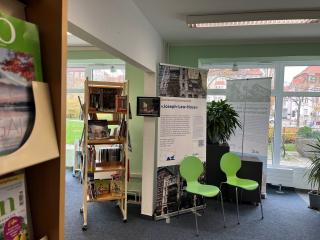 Zwei grüne Stühle vor einem Informationsdisplay mit Text und Bildern in einem hellen Raum mit großen Fenstern und Bücherregalen.