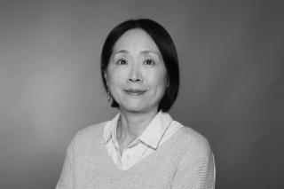 Person mit dunklen, schulterlangen Haaren trägt weißen Hemdkragen unter hellgrauem Pullover, vor einfarbigem Hintergrund.