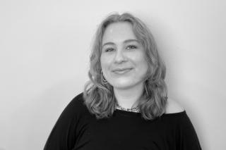 Person mit schulterlangen gewellten Haaren trägt ein schwarzes Oberteil mit Ausschnitt und eine Halskette vor hellem Hintergrund