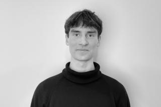 Person mit kurzem Haar trägt einen schwarzen Rollkragenpullover vor weißer Wand.