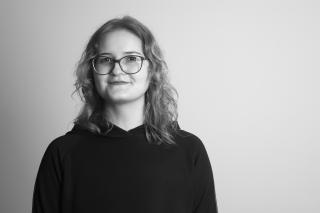 Person mit schulterlangen gewellten Haaren und Brille lächelt leicht in die Kamera und trägt einen dunklen Pullover.