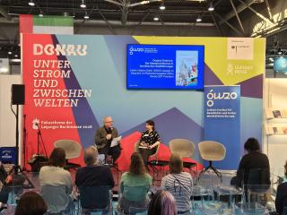 Zwei Personen sitzen auf Stühlen vor einer farbigen Messestandwand mit Texten und Logos, davor Publikum auf transparenten Stühlen in einer Messehalle.