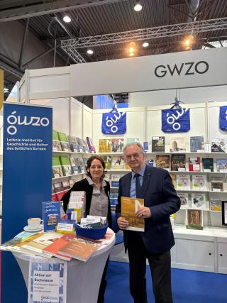Zwei Personen stehen an einem Messestand des Leibniz-Instituts für Geschichte und Kultur des östlichen Europa mit Büchern und Flyern auf einem Tisch und Regalen im Hintergrund, darüber ein Schild mit der Aufschrift 'GWZO'