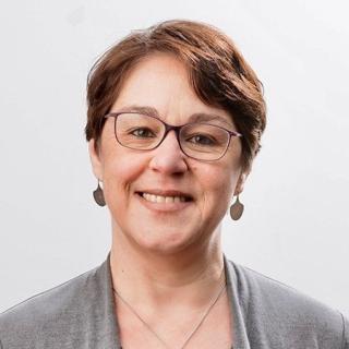 Portrait Manuela Boatca: Person mit kurzen braunen Haaren, grauer Strickjacke und silbernen Ohrringen und Halskette vor hellem Hintergrund.