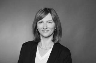 Frau mit schulterlangem, glattem Haar trägt ein schwarzes Sakko und ein weißes V-Ausschnitt-Oberteil vor einfarbigem Hintergrund.