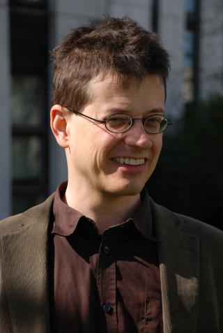 Mann mit kurzen, zerzausten Haaren und runder Brille trägt ein dunkles Hemd und ein braunes Jackett und lächelt im Freien.