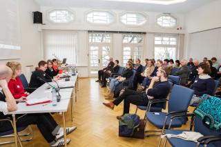 Großer, lichtdurchfluteter Tagungsraum, bei dem fast alle Sitzplätze belegt sind. Links ein Podium mit fünf Personen