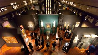 Blick auf die Ausstellung »Kaiser Karl IV. 1316-2016« in der Wallenstein Reithalle, Nationalgalerie Prag. Mehrere Vitrinen mit Kunstwerken, Besucher und zwei ausgestellte, beleuchtete Statuen im Zentrum.