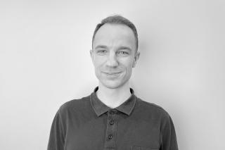 Person mit kurzen Haaren trägt ein dunkelgraues Poloshirt mit Knopfleiste vor weißer Wand.