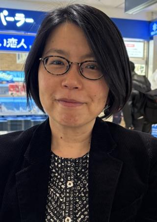 Frau mit schwarzem kurzem Haar, Brille und schwarzem Sakko vor einem Laden mit japanischer Beschilderung.
