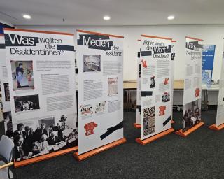 Fünf aufstellbare Roll-ups mit Text und historischen Schwarz-weiß-Fotos zur Dissidenz stehen in einem Raum auf grauem Teppichboden.