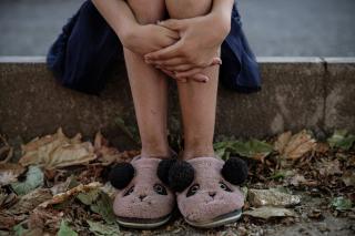 Die Beine eines Kindes sitzen auf einer Mauer, die Hände sind um die Knie gelegt, das Kind trägt Hausschuhe mit Tiergesichtern und Pompons, umgeben von trockenen Blättern.