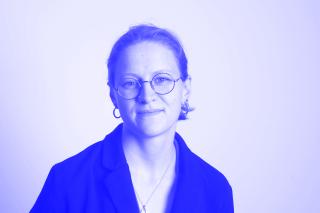 Schwarz-weiße Porträtaufnahme einer Person mit Brille, Ohrringen und zurückgebundenem Haar, die leicht lächelt.