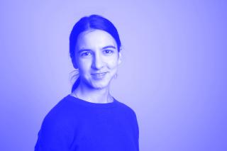 Porträt einer lächelnden Person mit dunklen Haaren, die zu einem Zopf gebunden sind, vor einfarbigem Hintergrund