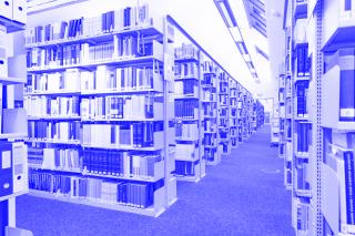 Ein langer Gang in der Bibliothek ist zu sehen, auf beiden Seiten zahlreiche Bücherregale, an der Decke eine Dachschräge mit Dachfenstern