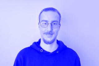 Mann mit zusammengebundenem Haar und Bart trägt schwarzen Kapuzenpullover vor weißem Hintergrund.