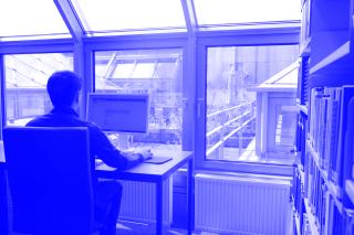 Person sitzt an einem Schreibtisch vor einem Computer in einem Raum mit großen Fenstern und Blick auf ein Dach mit Glasflächen.