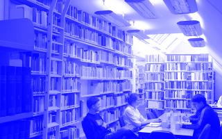 Vier Personen sitzen an einem Tisch in einem schmalen, hohen Raum mit Bücherregalen an den Wänden und Dachfenstern.