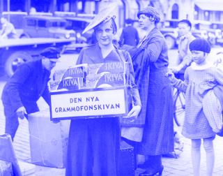 Schwarz-weiße Aufnahme einer Schallplattenverkäuferin in Stockholm der 1930er Jahre mit einem Schild 'DEN NYA GRAMMOFONSKIVAN' und mehreren Schallplatten in der Hand, im Hintergrund Passanten und Autos.