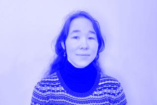 Person mit langen, gewellten Haaren trägt einen gemusterten Pullover über einem schwarzen Rollkragenpullover vor einfarbigem Hintergrund.