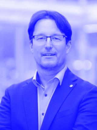 Mann mit Brille, dunklem Haar und blauem Sakko vor unscharfem Hintergrund.