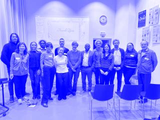 Skupinová fotografie účastníků a účastnic odborného setkání stojících v přednáškové místnosti. Za nimi je promítací plátno se snímkem „Thank You“, na stěně hodiny a informační panely. Lidé stojí v řadě, někteří s jmenovkami, v neformálním pracovním oblečení. Před nimi jsou prázdné židle.