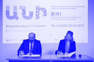 Zwei Personen sitzen an einem Tisch und unterschreiben Dokumente vor einer Wand mit einer Projektion zum Thema 'ANI The One Thousand and One Afterlives of a Medieval Armenian Capital' mit Ausstellungsdaten.