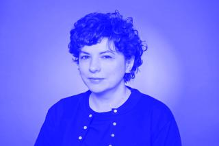 Person mit welligem, kurzem Haar, die ein schwarzes Oberteil mit Knöpfen trägt, vor einfarbigem Hintergrund