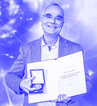 Můž stojící před modrým pozadím. Před sebou drží otevřené desky s diplomem a otevřenou krabičku s medailí.