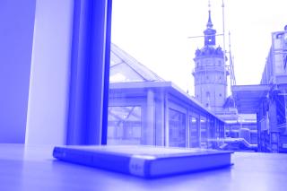 Ein Buch liegt auf einem Tisch an einer Fensterfront, hinter der man den Kirchturm der Nikolaikirche hervorragen sieht.