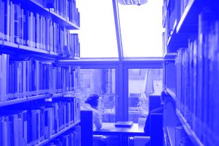 Am Ende eines Gangs mit Bücherregalen auf beiden Seiten sitzen zwei Personen an einer Fensterfront an Tischen und arbeiten am Laptop bzw. lesen in einem Buch.